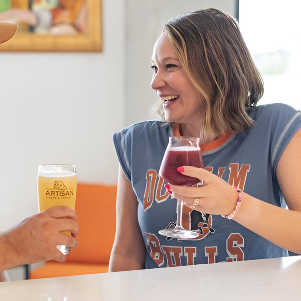 A smiling woman in a blue shirt holds a red drink, clinking glasses with another person.