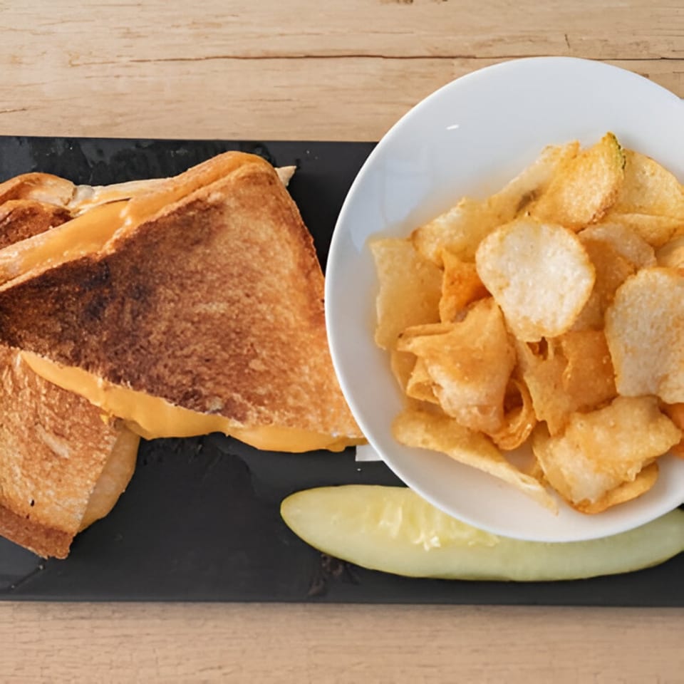Grilled cheese sandwich with a side of potato chips and a pickle spear on a black plate.