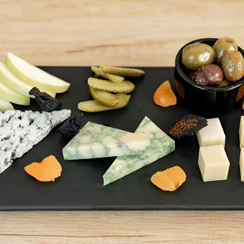 A black platter of Artisan Beer and Cheese's cheese, pickles, olives, dried fruit, and apple slices.