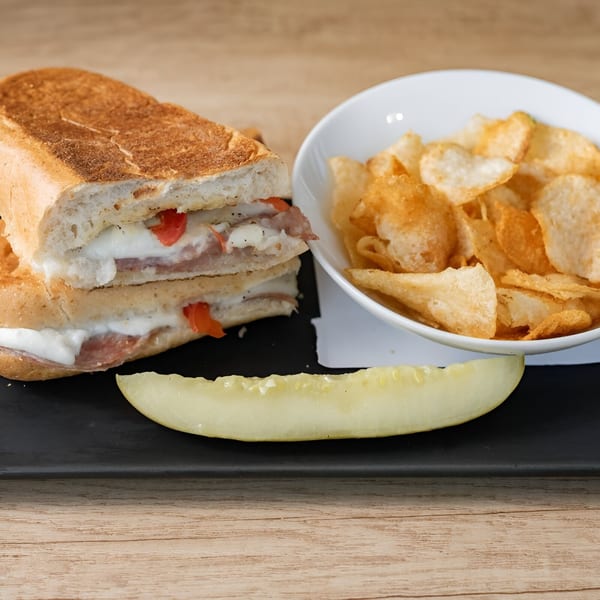 A panini with melted cheese and red peppers, a bowl of chips, and a pickle spear.